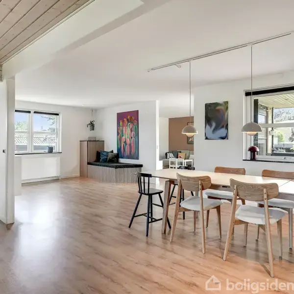 En moderne spisestue med et stort hvidt bord og seks stole. Et maleri og vinduer med udsigt til haven i baggrunden. Lys trælaminatgulv og loftslamper supplerer rummet.