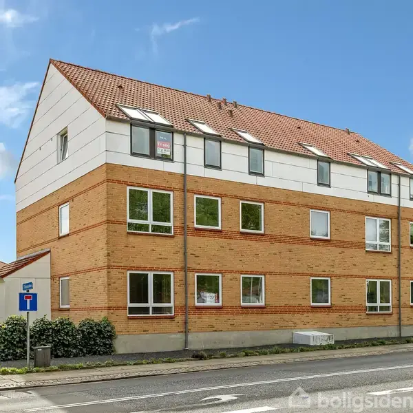 Et treetagers murstensbygning med hvide gavle står på en gadehjørne. Solen skinner, og omgivelserne inkluderer vejskilte, en fortov og grønt buskads.