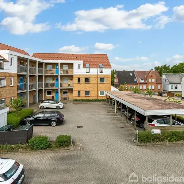 Lejlighedsbygning med rødtag og flere etager. Biler er parkeret i parkeringsområde med overdækket sektion til højre. Omgivelser inkluderer flere boliger og en blå himmel med skyer.