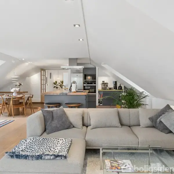 Et lyst og rummeligt loftsrum med skråvægge, hvor en grå sofa er centreret. I baggrunden ses et spisebord og et køkkenområde med moderne apparater og grønne planter.