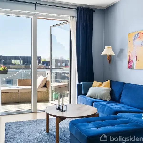 En blå sofa står i en lys stue ved siden af en åben altandør. En farverig kunstplakat hænger på væggen, og der en gulvlampe ved siden af sofaen.
