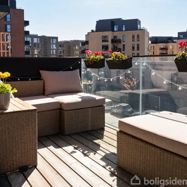 Terrassemøbler af rattan står på en solrig balkon med trægulv. Hængende blomsterkasser med røde og gule blomster pynter glasrækværket, mens lyskæder skaber en hyggelig atmosfære.