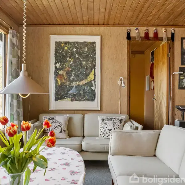 Sofa med lyse puder står i stue ved stort vindue; bord med dug og tulipaner foran. Malerier og dekorative figurer hænger på væg. Lys strømmer ind.
