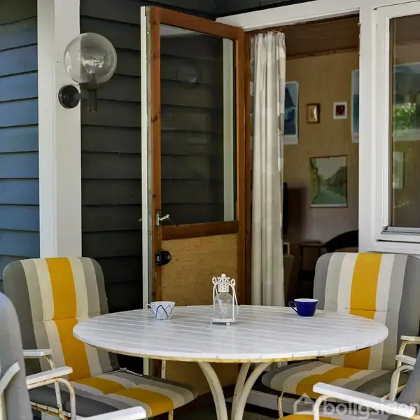 En rund, hvid havebord med fire stole står på en terrasse foran et hus med blå facade. En glasdør fører ind til en stue, hvor familiebilleder hænger på væggen.