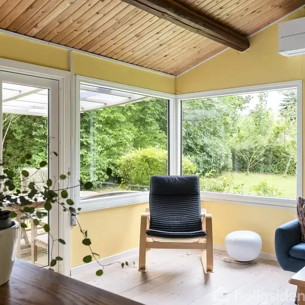 Stol står ved stort vindue, omgivet af en sofa og plante. Udenfor er der en grøn have med buske og træer, og rummet har gule vægge og træloft.