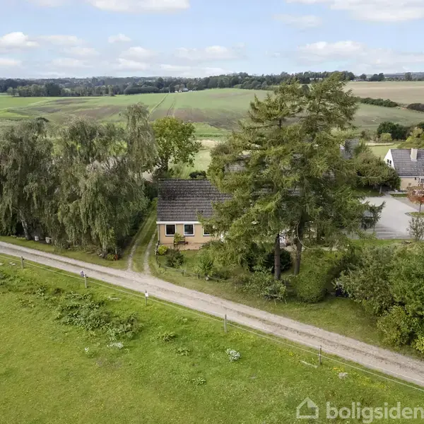 Hus står stille omgivet af grønne træer. Grusvej fører forbi huset i landlig omgivelser med marker og enkelte andre bygninger i baggrunden under en blå himmel.