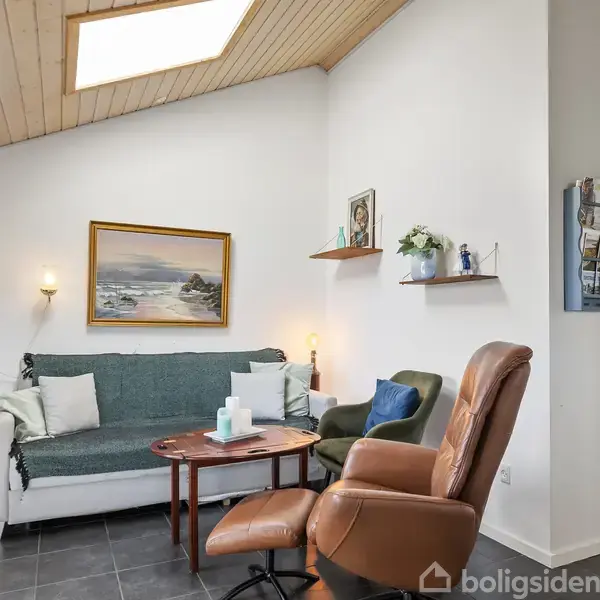 Et stueområde med en sofa og to lænestole ved et lille bord. Hvidmalede vægge prydet med kunst og hylder. En spiseplads og en åben dør i baggrunden.