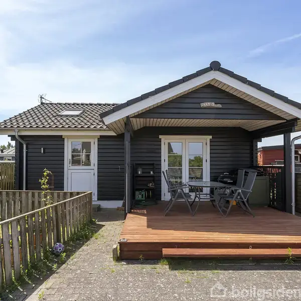 En stue med en sofa og lænestol, et maleri på væggen, et vægmonteret tv, og et sofabord. Rummet har lyse vægge, loft med ovenlys og flisegulv.
