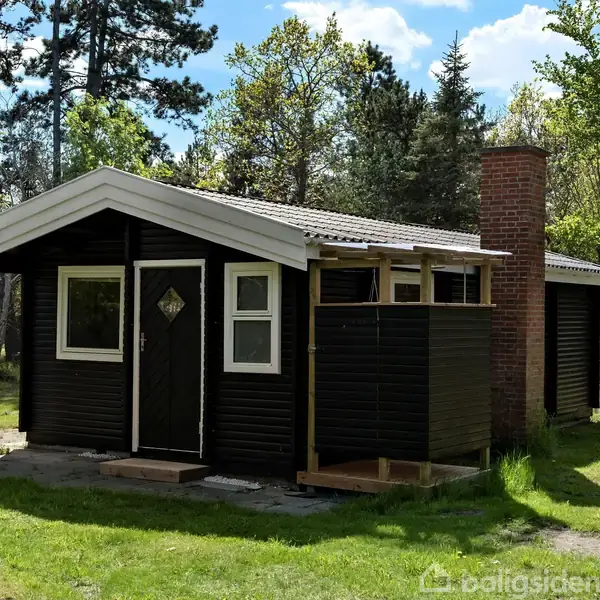 Et sort træsommerhus står stille i en skovklædt have. Huset har en hvidt tagudhæng, en skorsten og vinduer, der skuer ud mod omgivelserne af grønne træer og græs.