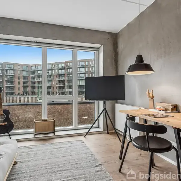 Et moderne værelse med en sofa til venstre, et skrivebord med lampe til højre, og en guitar samt fjernsyn nær vinduet. Der står "PIZZA PIZZA PIZZA" på en plakat.