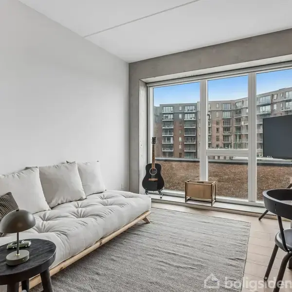 Sofa står i en lys stue foran store vinduer med udsigt til bygninger. En lampe er placeret på et lille bord ved siden af sofaen. En guitar står ved vinduet.