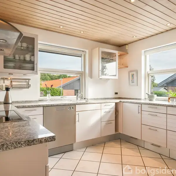 Et moderne køkken med hvid skabsplads og grå granitbordplader. Rummets vinduer slipper lys ind, og udenfor ses en fliseterrasse og grøn bevoksning. Kaffemaskine og en stålkande står på bordet.