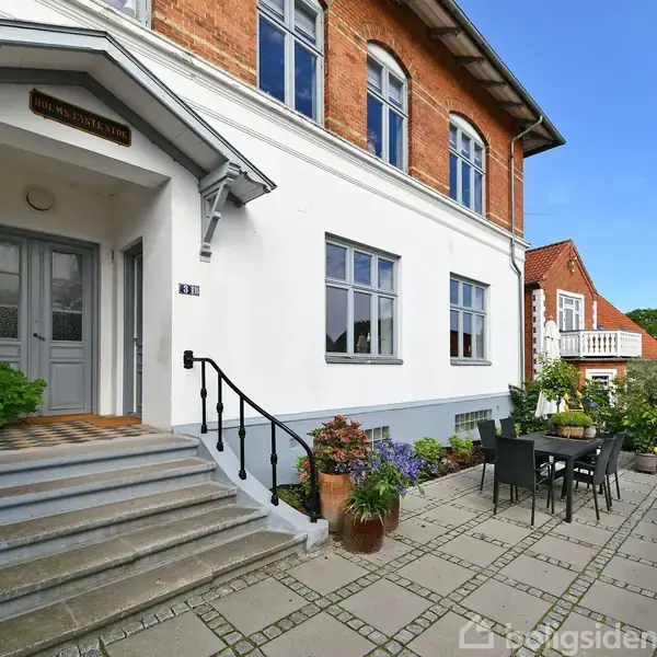 En trappe fører op til en hoveddør på en murstensbygning omgivet af en terrasse med borde, stole og planter. Tekst: "Helsingborg".
