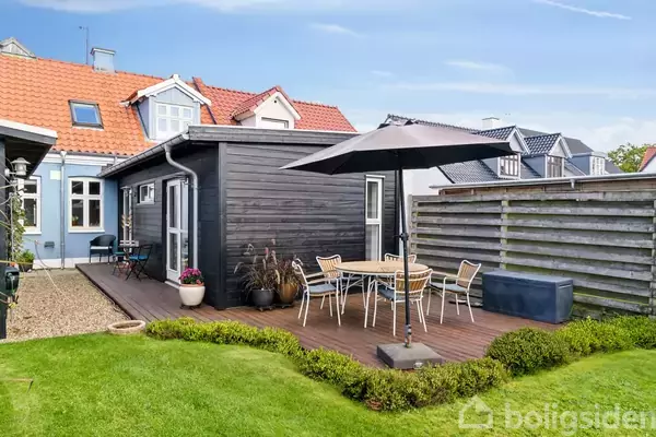 Sort træterrasse med hvidt cafébord og fire stole under stor sort parasol. Omgivet af grøn have og hegn, ved siden af et hus med blå og røde tage.