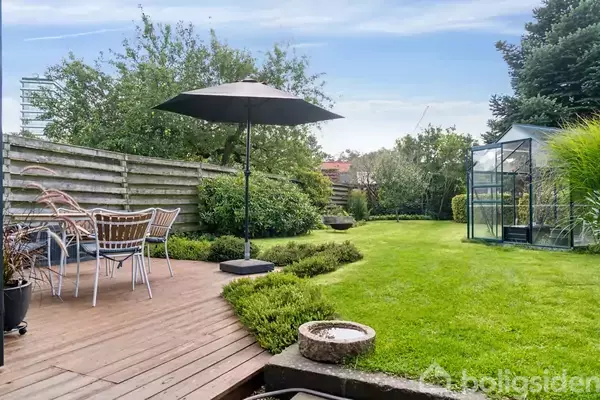 En parasol står på en træterrasse med stolesæt, omgivet af en velholdt græsplæne og buske, mens et drivhus er placeret i baggrunden nær et hegn.