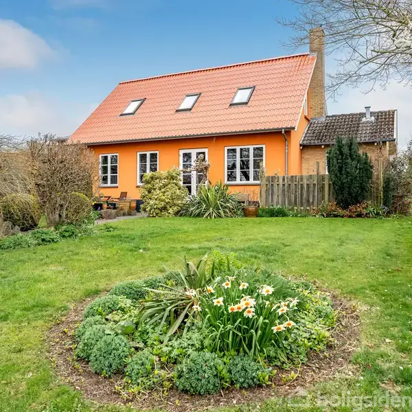 Et orange hus med rødt tegltag og tagvinduer står fredfyldt i en frodig have. Blomsterbede fyldt med hvide blomster omfavner huset, mens græsplænen breder sig foran.