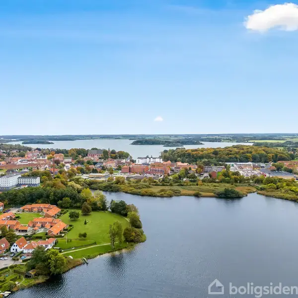 Bylandskab ses fra oven, hvor en stor sø strækker sig gennem det grønne område. Røde hustage og bygninger omringer søen, med skov og marker i baggrunden under en klar himmel.