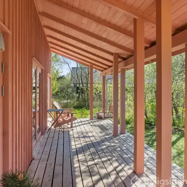 Veranda med lyserøde trævægge og loft, adskilte af træstolper, med udsigt til en have. En dør har en rund rude. I baggrunden ses et træhus.