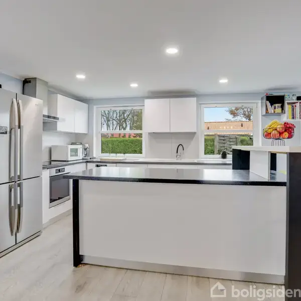 Et moderne køkken med en stor, hvid ø, omgivet af skabe og hårde hvidevarer som et køleskab, ovn og mikrobølgeovn. Vinduer giver udsigt til en grøn have.