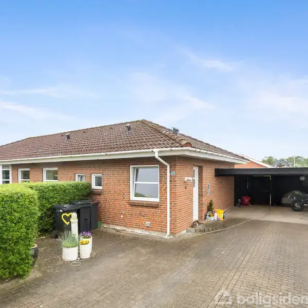 Rødt murstenshus med skråt tag, veranda med carport, og indkørsel i forgrunden. To affaldsbeholdere og blomsterkrukker ved siden af en klippet hæk. Klar blå himmel.
