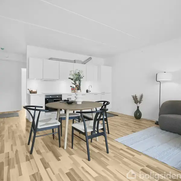 Et moderne køkken-alrum med et rundt spisebord og fire stole i forgrunden. En sofa og gulvlampe står til højre. Lyse trægulve og minimalistisk indretning præger rummet.