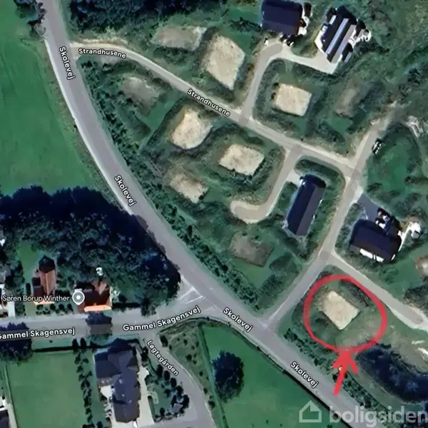 Luftfoto viser et boligområde med markerede byggegrunde. En rød pil peger mod en specifik grund. Vejnavne inkluderer "Gammel Skagensvej" og "Strandholmsvej" omgivet af græsarealer og træer.