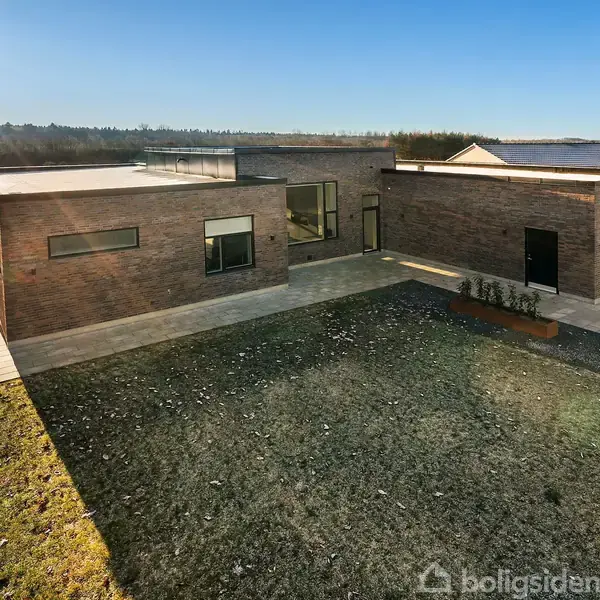 Et moderne hus står stille på en flisebelagt terrasse i en åben have med græsplæne. I baggrunden ses en skovlinje under en klar, blå himmel.