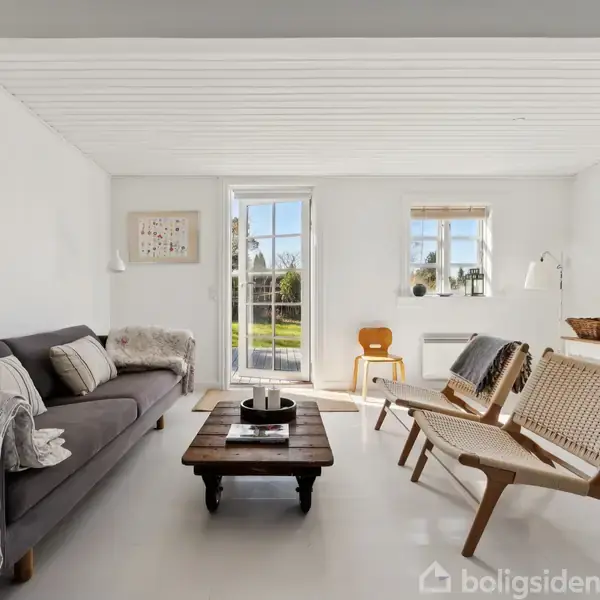 En lys stue med en grå sofa til venstre dækket med tæpper og puder. Et træbord står foran sofaen. Til højre står to stole. I baggrunden er der vinduer og en dør, der åbner mod en grøn have.