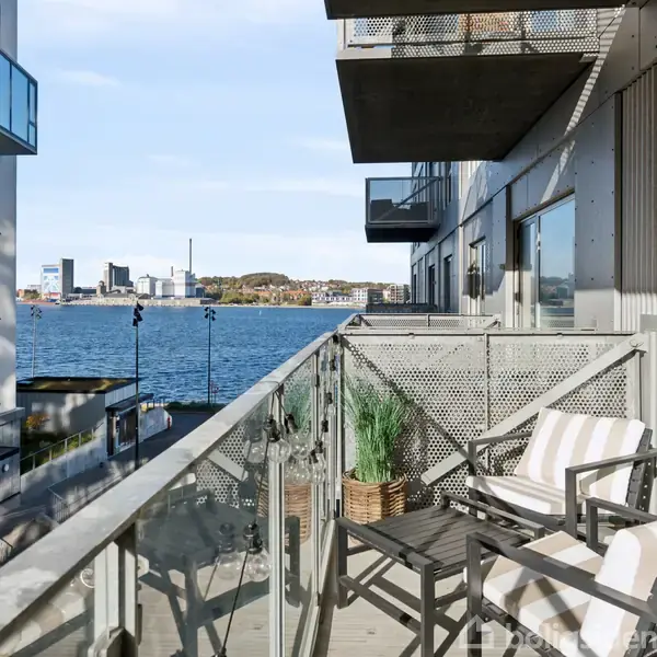 To stole står med hynder på en altan, flankeret af en plantekasse. Udsigt over vandet med moderne bygninger i baggrunden under en klar himmel.