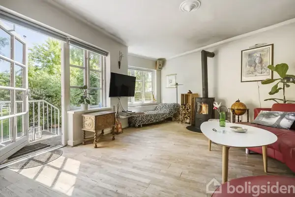 Stue med træbrændeovn i hjørnet, rød sofa og rund hvid sofabord. Fladskærms-tv monteret på væggen, zebrastribet daybed og vinduer åbner til en grøn have udenfor.