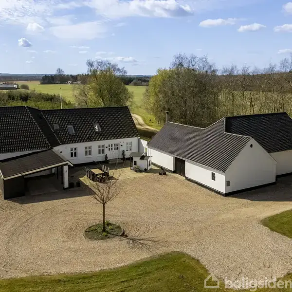 Et hvidt hus med mørkt tag står på en grusplads omgivet af græs og træer. Det ligger i et landligt område med marker i baggrunden.