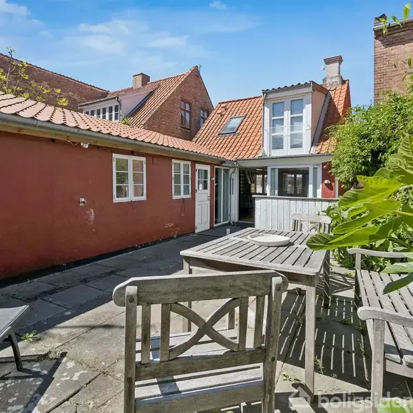 Et gårdhavemiljø med træbord og stole står på en flisebelagt terrasse. Bagved ses røde murstensbygninger med rødt tegltag, under en klar blå himmel.