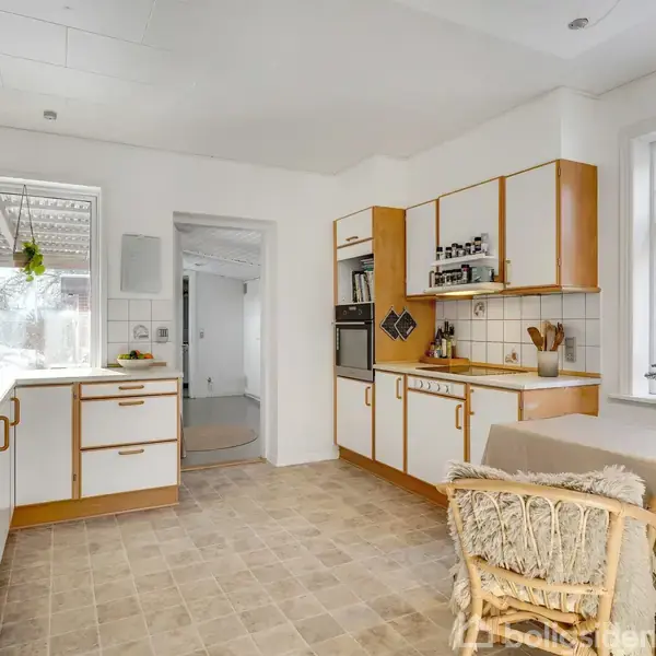 Et lyst køkken domineret af hvide skabe med trækanter og en beige flisegulv. Et spisebord med fletstol står ved vinduet, omgivet af lysindfald og grønne planter.