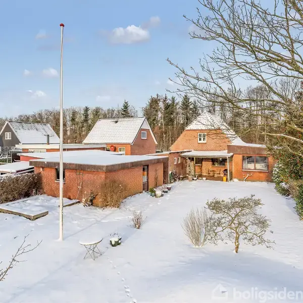 En rød murstenshus står i en snedækket have med træer omkring. Der er en flagstang foran huset, og nabohuse ses i baggrunden under en blå, skyet himmel.