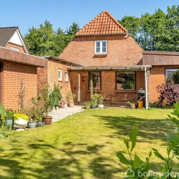 Et rødt murstenshus med en lille kvist og rødt tegltag. Foran huset er der en velplejet græsplæne med forskellige planter i potter. Bag huset ses grønne træer.