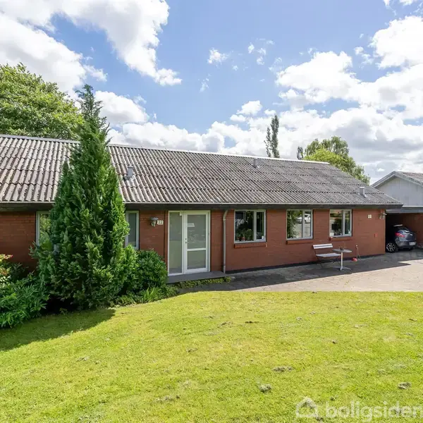 En murstensvilla med skråtag står på en grøn græsplæne omgivet af træer. Ved indgangen er en bænk, og en indkørsel fører til en carport. Himmel med skyer.