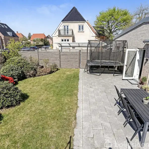 Trampolin står stille i en have med grøn græsplæne og farverige blomsterbuske; terrasse med spisebord og stole ved siden af huset i et boligkvarter.