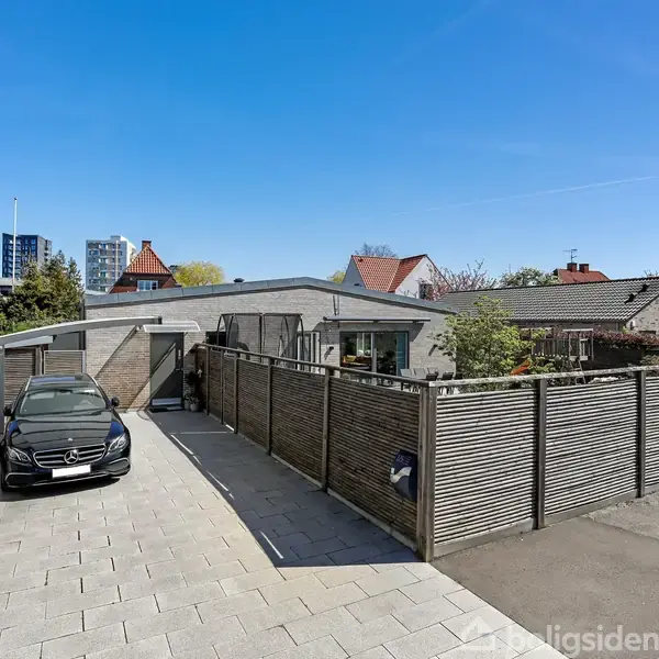 En sort bil parkerer på en flisebelagt indkørsel ved siden af en moderne enetages bolig med plankeværk. I baggrunden ses flere huse og nogle træer under en klar blå himmel.