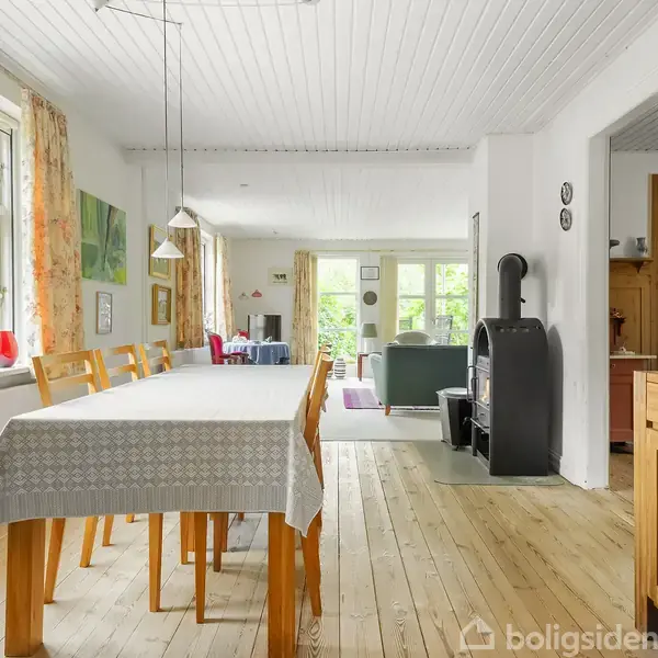 Spisebord med dug og stole står i lys, rummelig stue med trægulv. Stuen har vinduer og dør ud til haven, en brændeovn står i hjørnet.