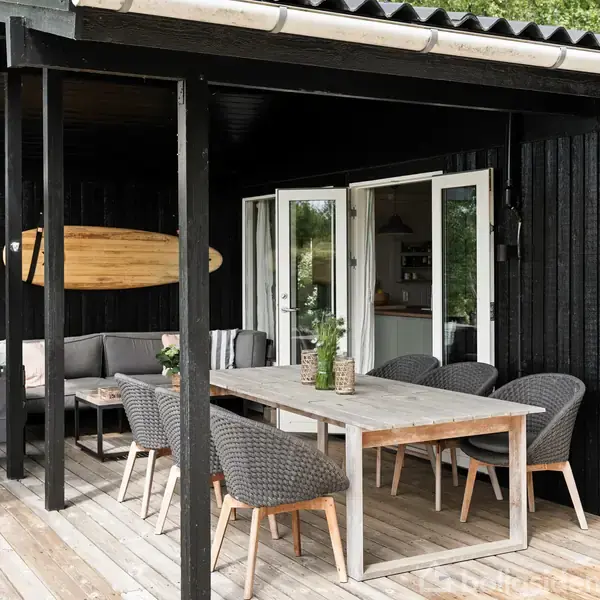 Et træbord omgivet af flettede stole står på en terrasse. En sofa står ved siden af under et surfbræt på væggen. Døre fører ind til et køkken.