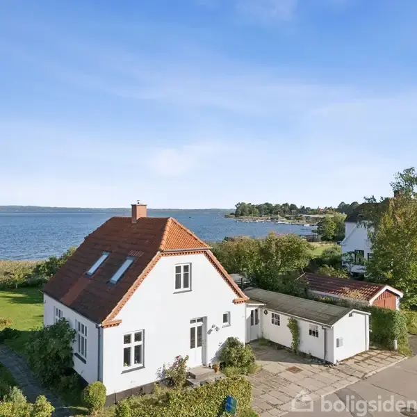 Hvidt hus med rødt tag står stille i en naturskøn kystnær område, omkranset af grønt græs og skov, med udsigt til en rolig blå sø.