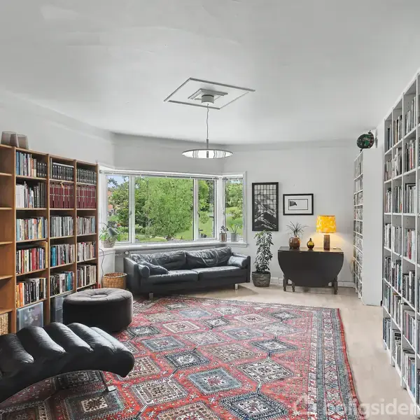 Stue med lædersofa og chaiselong på et flerfarvet tæppe, omgivet af bogreoler. Store vinduer bag sofaen giver udsigt til grønt. Et bord med lampe står i hjørnet.