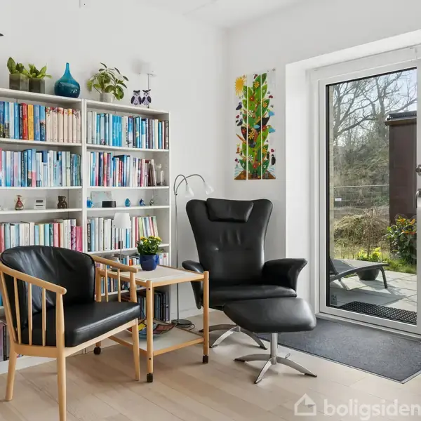 Bogreol fyldt med bøger og dekorationer står i en lys stue. Ved siden af står to stole og et lille bord. Store glasdøre åbner ud til en terrasse med udsigt til haven.
