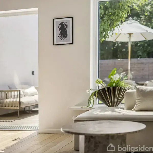 Stue med sofa og sofabord, åbne døre fører til terrasse med parasol. Indretning inkluderer grønne planter og minimalistisk kunst på væggen.
