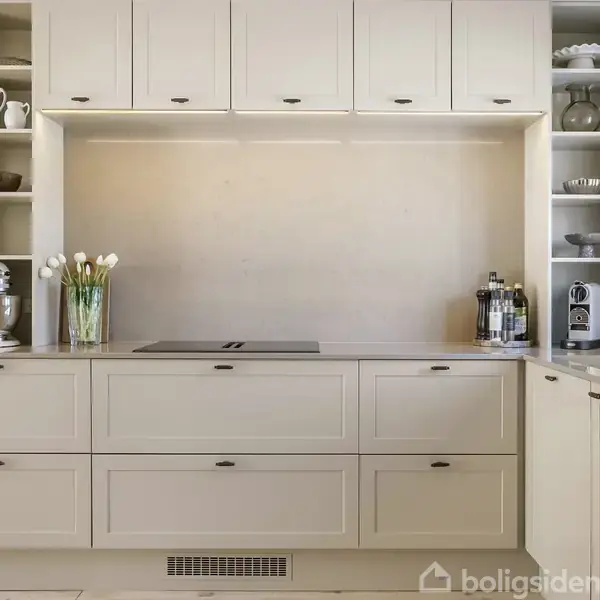 Et moderne køkken med beige skabe indeholder en kogeplade, blomster i en vase og køkkenudstyr. Hylder på begge sider viser porcelæn i en lys, indbydende atmosfære.