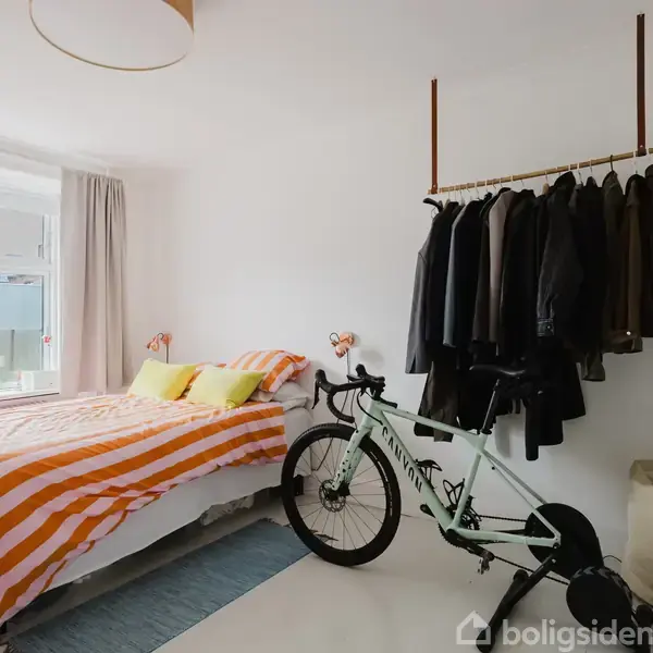 En cykel står på en gulvmåtte ved siden af en seng med orange stribede dyner. Jakker hænger på et stativ over cyklen. Rummet er lyst med et stort vindue.