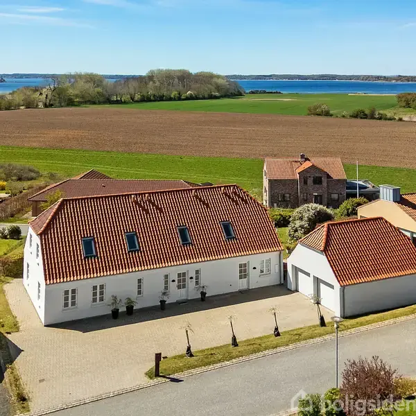 Hvidt hus med rødt tegltag står stille ved siden af en mindre garage. Det ligger på en brolagt vej omgivet af marker, huse og udsigt til vand i baggrunden.
