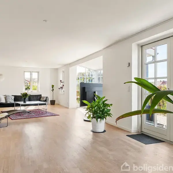 Stue med sofa og lænestol, indrettet med tæppe og planter, vendt mod glasdøre, der leder til have. Lyse trægulve og minimalistisk indretning skaber et rummeligt udseende.
