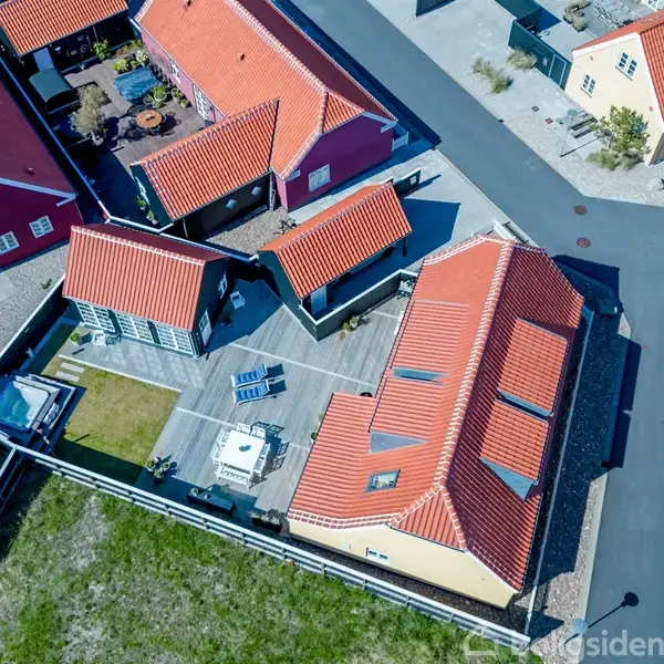 Huse med røde tegltage set ovenfra ligger på en stille vej. Et åbent gårdrum med møbler er placeret mellem bygningerne, omgivet af en grøn græsplæne og grusbelagte omgivelser.