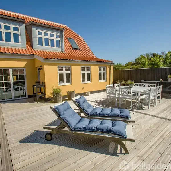To liggestole med blå puder står på en stor træterrasse ved et gulmalet hus med rødt tag. Ved siden af står et hvidt havebord. Skov ses i baggrunden.